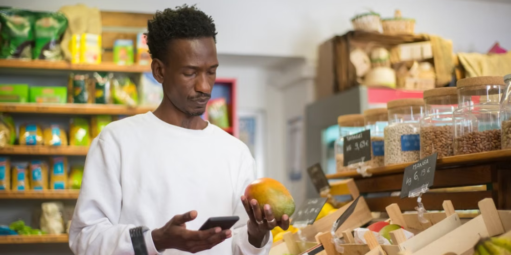 Homem negro de camiseta branca segura um celular e uma manga no mercado